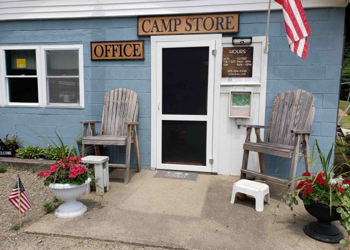 Entrance to a camp store and office with two wooden chairs, flower pots, an American flag, a posted sign with hours, and a small white stool by the door.