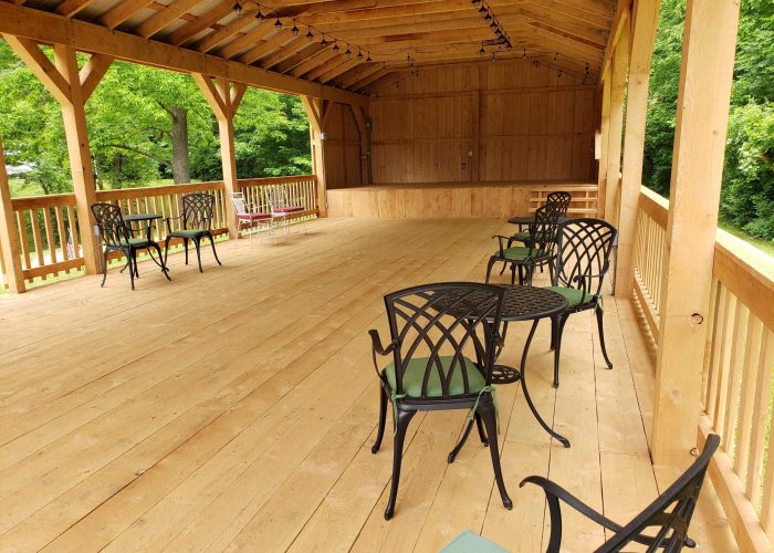 Covered wooden pavilion with several black metal tables and chairs spaced apart on the wooden floor; green trees visible through the open sides.